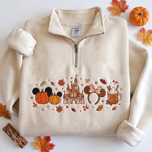 Op de afbeelding: Een crèmekleurige sweater met een korte rits en een geborduurd ontwerp van twee pompoenen met muizenoortjes, een kasteel, muizenoortjes met een geruite strik en een mok warme chocolademelk, omringd door kleine herfstblaadjes en besjes.