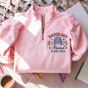 以下が含まれることがあります： 左胸に刺繍が施されたライトピンクのクォータージップスウェットシャツ。デザインには、青い肋骨のイラスト、花のアクセント、そしてダークブラウンとブルーの文字で「RADIOLOGY Mama X-RAY TECH」というテキストが含まれています。