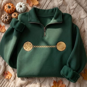 May include: A dark green quarter-zip sweatshirt featuring an embroidered gold design across the chest. The embroidery consists of a compass rose on the left, a circular geometric emblem on the right, and a thin chain connecting the two symbols.