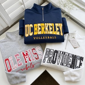 May include: Three quarter-zip sweatshirts featuring collegiate-style applique lettering. The navy sweatshirt displays yellow UC BERKELEY VOLLEYBALL text, the gray one shows red OLE MISS rebels text, and the white one features black PROVIDENCE text.