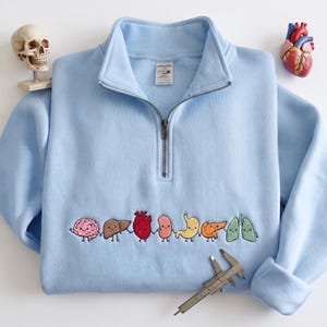 Puede incluir: Una sudadera azul claro con cremallera corta que presenta un bordado de órganos humanos de dibujos animados tomados de la mano, incluyendo cerebro, hígado, corazón, riñón, estómago, páncreas y pulmones. Un calibrador metálico está colocado.