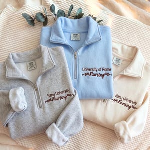 May include: Three quarter-zip sweatshirts in light blue, heather gray, and cream. Each features embroidered text: the blue one reads "University of Rome Nursing," while the others read "Hanu University Nursing" in dark brown with small bow accents.