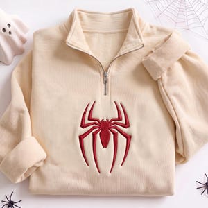 Può includere: Una felpa color crema con mezza zip e un vistoso emblema a forma di ragno ricamato in rosso al centro del petto. Il capo presenta un collo alto con chiusura a zip in metallo argentato e maniche lunghe con polsini a coste.