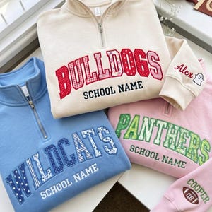May include: Three quarter-zip sweatshirts in cream, light blue, and light pink. The cream sweatshirt features red patterned letters reading BULLDOGS and SCHOOL NAME, with Alex and a ghost embroidered on the sleeve. The blue one says WILDCATS and SCHOOL NAME. The pink one says PANTHERS and SCHOOL NAME, with COOPER and a football on the sleeve.