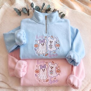 May include: A light blue quarter-zip sweatshirt and a light pink sweatshirt, both featuring an embroidered design of two ghost characters, pumpkins, and candy with the text "hey boo" in purple thread, centered on the front of each garment.