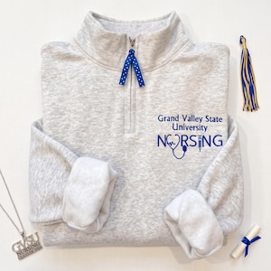 Puede incluir: Un suéter gris claro jaspeado con cremallera corta que presenta un diseño bordado en azul con el texto Grand Valley State University NURSING, junto a un estetoscopio y una jeringa. Una cinta azul con puntos blancos cuelga del tirador.
