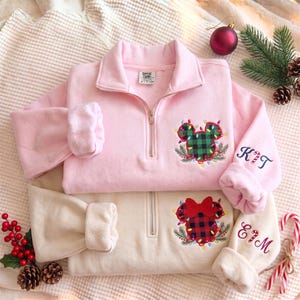 Puede incluir: Dos sudaderas con cremallera de un cuarto en rosa claro y crema. La sudadera rosa presenta una cabeza de Mickey a cuadros verdes con luces navideñas y el nombre KIT en la manga. La sudadera crema tiene una cabeza de Minnie a cuadros rojos con un lazo rojo, luces navideñas y el nombre EIM en la manga.