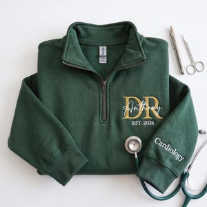 May include: A dark green quarter-zip sweatshirt featuring gold embroidered letters DR, the name Anthony in white script, and the text EST. 2026. The left sleeve is embroidered with the word Cardiology in white. A stethoscope and medical tools are nearby.