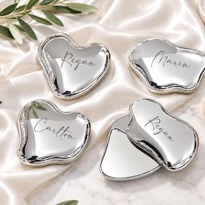 Peut inclure: Quatre miroirs de poche argentés en forme de cœur irrégulier avec une finition polie et réfléchissante. Chaque miroir présente un nom personnalisé gravé dans une police cursive noire, incluant Regan, Marin et Carlton, avec un miroir ouvert.