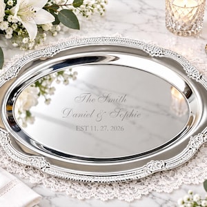 Puede incluir: Una bandeja de servicio ovalada de color plateado con un borde ornamentado y en relieve. El centro es pulido y reflectante, con el texto grabado: The Smith, Daniel & Sophie, EST 11. 27. 2026. La bandeja tiene un acabado metálico liso.
