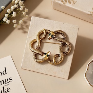 May include: A gold-toned metal brooch featuring two intertwined snakes forming a circular shape. Each snake head is accented with a small green gemstone eye. The brooch is displayed on a square, light-coloured textured card, showcasing detailed scale patterns along the bodies of the snakes.