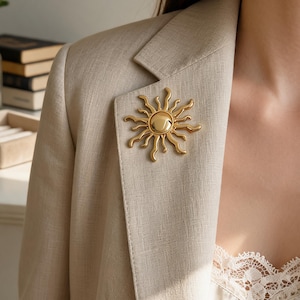 May include: A gold-toned sun-shaped brooch featuring a smooth, rounded centre and radiating, wavy rays. The accessory is pinned to the lapel of a beige blazer, which is worn over a cream-coloured camisole with delicate lace trim along the neckline.