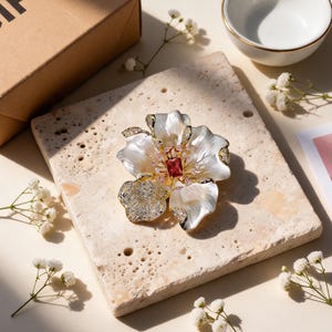 May include: A flower-shaped brooch featuring iridescent white petals with gold-toned edges, a central rectangular red gemstone, and small clear crystals. The brooch has a metallic gold-colored base and is displayed on a textured stone surface.