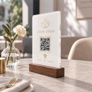 May include: A vertical clear acrylic sign holder with a white insert featuring a gold logo, a QR code, a Wi-Fi symbol, and the text "YOUR LOGO" and "SCAN TAP ENJOY". The sign is mounted in a rectangular dark brown wooden base on a table.
