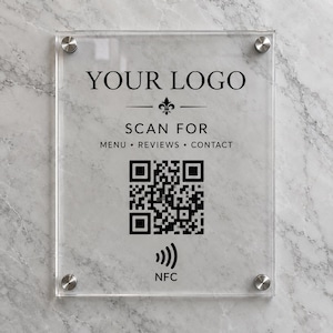 以下が含まれることがあります： 透明なアクリル製の長方形の看板。黒い文字で「YOUR LOGO」「SCAN FOR」「MENU」「REVIEWS」「CONTACT」「NFC」と記され、中央にQRコードとフルール・ド・リスのアイコンが配置されています。四隅は銀色の金属製スタンドオフで固定されています。