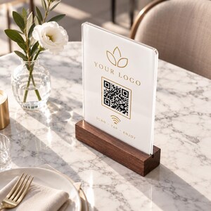 Könnte beinhalten: Ein transparenter Acryl-Schildhalter mit einem rechteckigen Sockel aus dunklem Holz. Das Schild zeigt ein goldenes Logo-Symbol, den Text "YOUR LOGO", einen zentralen QR-Code und den Text "SCAN - TAP - ENJOY" in goldener Schrift.
