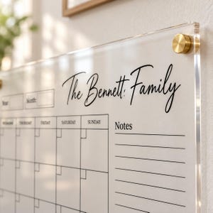 Könnte beinhalten: Ein Wandkalender aus klarem Acryl mit dem Text The Bennett Family in schwarzer Schreibschrift. Die Tafel enthält ein Raster für die Wochentage, Felder für Jahr und Monat sowie einen Notizbereich. Befestigt mit goldfarbenen, runden Halterungen.