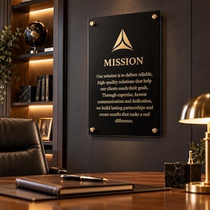 以下が含まれることがあります： 金色の三角形のロゴと「MISSION」という文字、それに続く信頼性の高いソリューションの提供に関する文章が記された黒いアクリルの長方形の壁掛け看板。四隅は金色の円形スタンドオフで固定されています。
