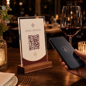 May include: A vertical acrylic sign holder with a wooden base displaying a DASE DESIGN logo, a QR code, and a contactless payment symbol. The sign is positioned next to a smartphone showing a matching contactless icon and a glass of red wine.