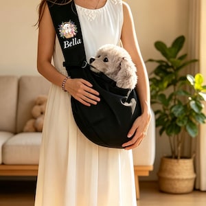 以下が含まれることがあります： 花かごに入った小さな白い犬のグラフィックと「Bella」という名前がプリントされた黒いペット用スリングバッグ。胸元に斜め掛けするタイプで、中にはふわふわとした明るい毛色の犬が顔を出しています。