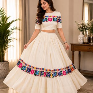 May include: A two-piece off-white outfit consisting of a cropped top and a long, pleated skirt. Both pieces feature horizontal bands of vibrant, multi-colored floral embroidery in shades of pink, blue, green, and yellow across the fabric.