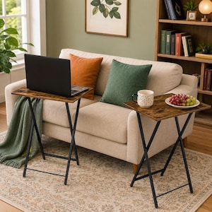 May include: Two folding snack tables with dark brown wood-grain laminate tops and black metal X-frame legs. One table holds a black laptop, while the other holds a white patterned mug and a white plate filled with red and green grapes.