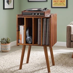 May include: A wooden record player stand with four legs, featuring three vertical storage compartments. The top surface holds a black record player. The compartments contain several books, a pair of grey over-ear headphones, and a collection of vinyl records.