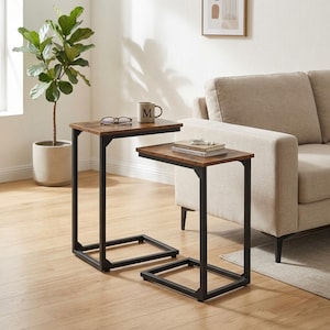 May include: Two C-shaped side tables with dark brown wood-grain tops and black metal frames. The taller table holds a gray mug with the letter M and a pair of glasses. The shorter table holds a stack of two books with a small object on top.