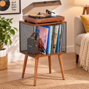 May include: A wooden record player stand with four tapered legs, featuring a black metal mesh side panel and an open storage compartment filled with vinyl records and a small brown portable speaker with the Marshall logo on the front.