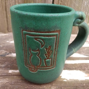May include: A green ceramic mug with a brown silhouette of a cat and a plant in a pot inside a frame. The mug has a handle that is shaped like a cat's ear.