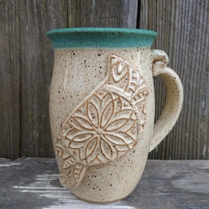 Puede incluir: Una taza de cerámica con un esmalte beige moteado y un borde turquesa. La taza tiene un diseño en relieve de una flor estilizada en el lateral. La taza tiene un asa con forma de flor estilizada.
