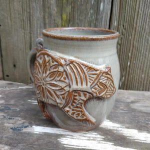 May include: A ceramic mug with a brown bear design. The bear is carved into the side of the mug and has a detailed pattern.
