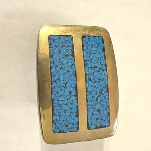 May include: A rectangular brass belt buckle with rounded corners featuring two vertical channels filled with small, crushed bright turquoise blue stones. The metal surface has a polished, reflective gold-tone finish surrounding the inlaid stone sections.