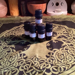 May include: Five small brown glass bottles with labels that read "Blessed Altar Oil 5ml". The bottles are arranged in a circle on a black and green fabric with a tree of life design.