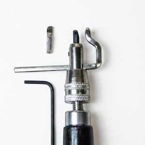 Peut inclure: Un outil de rainage pour cuir en métal argenté avec un manche noir, comprenant un guide réglable, une petite lame interchangeable et une clé hexagonale noire en forme de L. L'outil possède une section moletée pour le réglage de la lame.