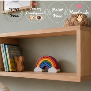 May include: A wooden floating shelf holding a stack of books, a small wooden mouse-shaped figure, and a rainbow-shaped toy with white cloud bases. Text overlays indicate the product is BPA Free, for sensory development, paint free, and handmade.