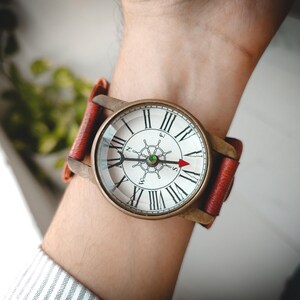 May include: A round wristwatch with a bronze-toned metal case and a red leather strap. The white dial features black Roman numerals, a central ship wheel graphic, and a red arrow-tipped hand pointing towards the letter S on the dial face.