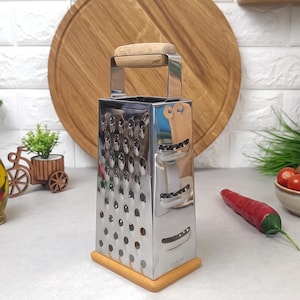 Puede incluir: Un rallador de caja de cuatro lados hecho de acero inoxidable con un mango de madera de color claro en la parte superior y una base de madera a juego. El rallador presenta varias superficies de corte perforadas en sus lados para rallar y rebanar alimentos.