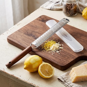 Puede incluir: Un rallador de cítricos de acero inoxidable con mango de madera de color marrón oscuro. La herramienta cuenta con una hoja de metal larga y estrecha con pequeñas perforaciones cuadradas afiladas. Incluye una funda protectora de plástico.
