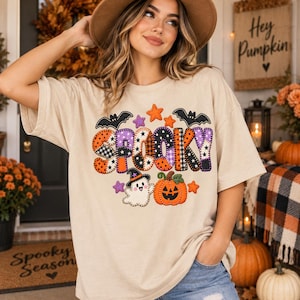 May include: A beige short-sleeve t-shirt featuring a colorful graphic with the word SPOOKY in patterned block letters, surrounded by illustrations of two black bats, orange and purple stars, a white ghost wearing a hat, and an orange pumpkin.