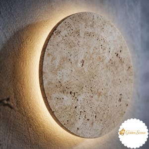 May include: A circular wall sconce made of beige, porous stone with a natural textured surface. The fixture emits a warm, soft glow from behind its edges, creating a halo effect against the wall. The logo Golden Sconce is visible in the corner.