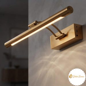 May include: A brushed gold wall-mounted picture light with a long, cylindrical horizontal lamp head. The fixture features a rectangular base plate and two adjustable curved arms connecting the base to the light bar, emitting a warm glow.