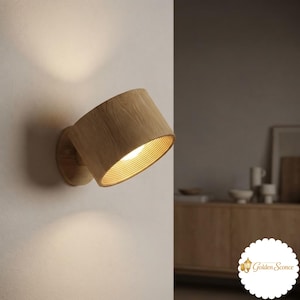 Könnte beinhalten: Eine zylindrische Wandleuchte aus hellem Holz mit sichtbarer Maserung. Die Leuchte ist an einer Wand montiert und verfügt über eine abgewinkelte Öffnung, die ein warmes, helles Licht abgibt und die umliegende Wandfläche beleuchtet.
