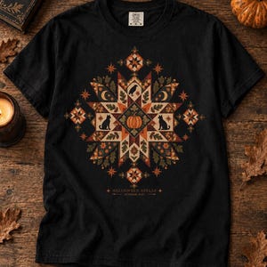 May include: A black short-sleeved t-shirt featuring a large, intricate, geometric quilt-style graphic in shades of orange, brown, and cream. The design includes a central pumpkin, black cats, a raven, moons, and leaves, with the text HALLOWEEN SPELLS OCTOBER 31ST.