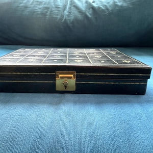 May include: A rectangular black storage box with a hinged lid featuring a grid pattern of small gold-colored symbols. The box has a gold-colored metal latch with a keyhole on the front edge and a thin gold-colored decorative border along the lid.