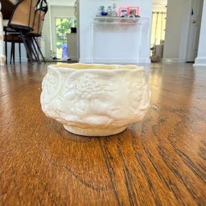 May include: A cream-colored ceramic bowl featuring a raised, embossed floral pattern around the exterior. The bowl has a scalloped rim and a rounded, squat shape, with a glossy finish that reflects light across its textured, sculpted surface.