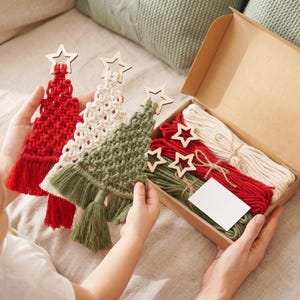 May include: Three macrame Christmas tree ornaments in red, cream, and olive green, each topped with a wooden star. A cardboard box contains additional coiled red, cream, and olive green cord, three wooden stars, and a blank white rectangular card.