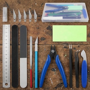 May include: A model kit tool set including a 15 cm metal ruler, five replacement blades, a clear storage case, a green polishing cloth, a hobby knife, blue side cutters, two pairs of black tweezers, a metal file, and two sanding sticks.