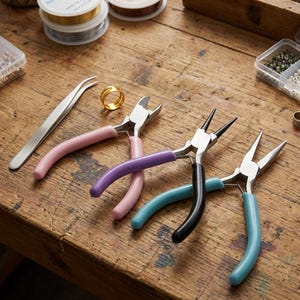 May include: A set of jewelry making tools including a pair of silver tweezers, a gold ring, and three pairs of pliers with pink, purple, black, and light blue rubberized handles, arranged on a wooden surface with spools of wire and beads.