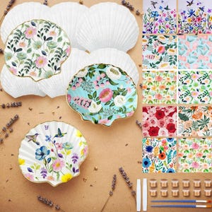 Può includere: Un set di piatti in ceramica a forma di conchiglia con vari motivi floreali e di colibrì. La collezione include conchiglie bianche semplici, conchiglie dipinte con disegni botanici colorati, piccoli vasetti di vernice e due pennelli sottili.